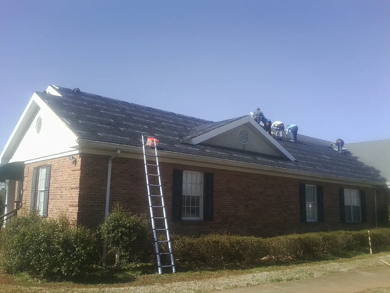Crew tearing shingles off a brick ranch roof with a ladder set — Emergency Roof Repair work by our crew in Baldwinville