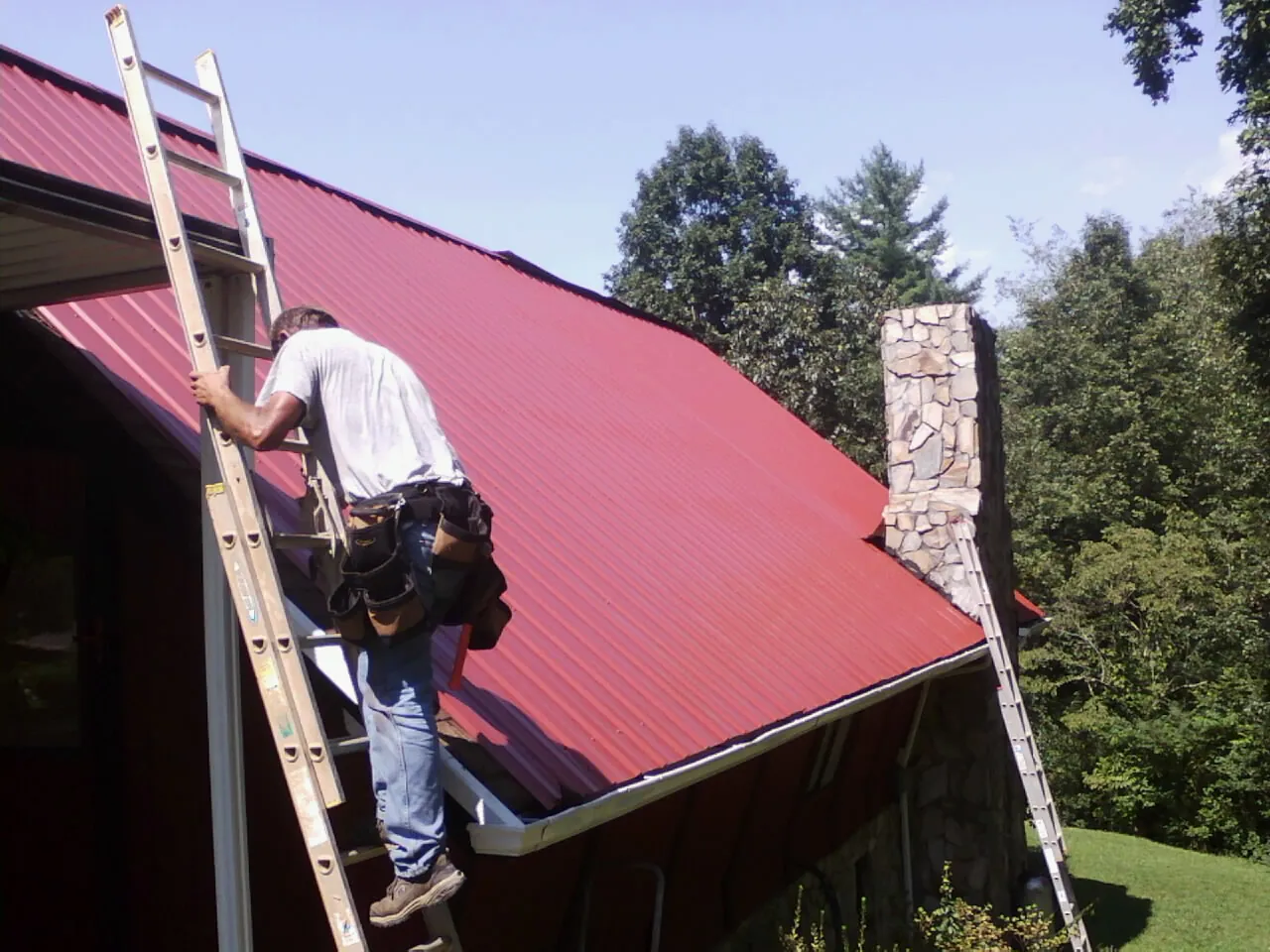 Roofer on a ladder beside a red metal roof and stone chimney — Soffit & Fascia Repair by Roofing Brothers in Baldwinville